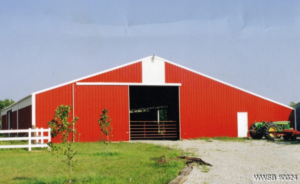 A large red barn with a white roof, reminiscent of modern agricultural steel buildings, sits under a clear blue sky. The barn has an open door revealing farm equipment inside. A tractor is parked nearby on gravel, with a few small trees and a patch of grass in the foreground.