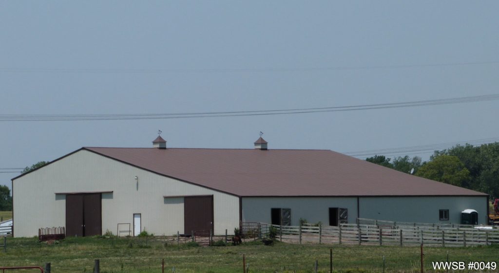 In a rural landscape, this large agricultural steel building features a brown roof with two small cupolas. Surrounded by fencing, it stands amidst grass in the foreground and a few trees under a clear sky.