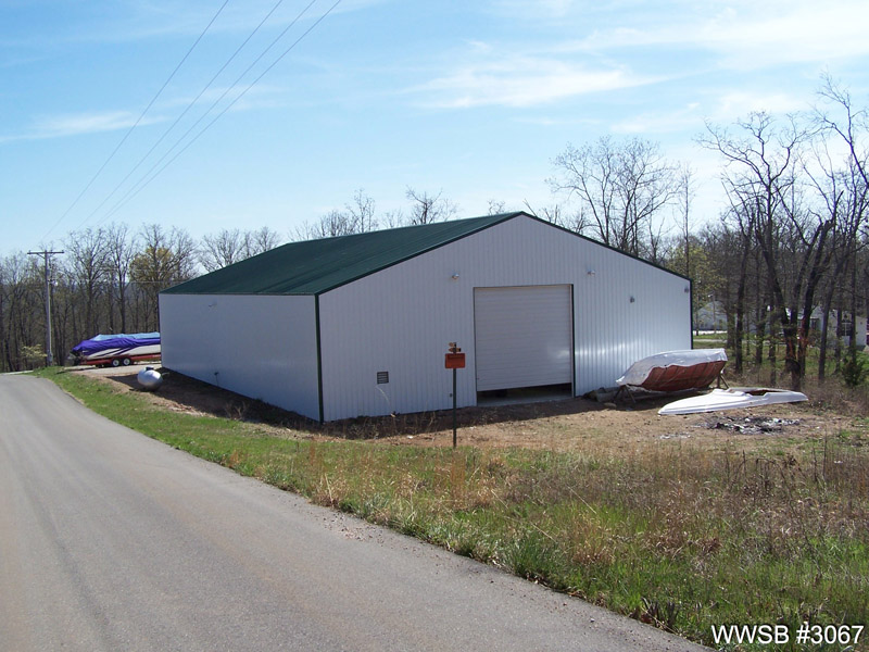 A large commercial metal building with a green roof and white walls under a clear blue sky. There's a large garage door on the front side and a standard entry door beside it. To the left, there's a boat covered with a blue tarp, and some debris is scattered on the ground nearby. The building is situated beside a paved road with power lines overhead, and a wooded area can be seen in the background.