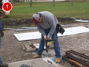 A construction worker in a gray sweater and cap uses a power drill on a concrete surface outdoors, contributing to steel building construction. Tools and metal sheets are scattered nearby on the gravel area.