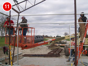 Construction workers are assembling a steel framework, with some standing on a cherry picker while others work on a platform. The site is filled with tools, beams, and a vehicle in the background. The number 11 is visible in the top left corner.