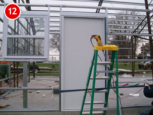 Construction of a steel building with a metal frame featuring a window and door partially installed. A green ladder with a yellow top holds a power drill. The number 12 is in a red circle on the top left. Vehicles and trees are visible in the background.