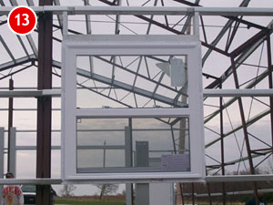 A white, rectangular window frame is set within a steel framework of a greenhouse under construction. The sky is cloudy, and a red circle with the number 13 is in the upper left corner.