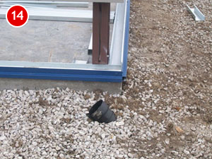 Partial view of a steel building foundation with blue insulation along the edge. A black pipe protrudes from crushed gravel next to the foundation. The surroundings appear to be under construction or landscaping.