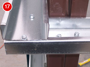 Close-up of a metal construction corner with overlapping steel panels, indicative of steel building construction, fastened by bolts. The panels are silver and brown, showing a precise fit and finish. A red circle with the number 17 is visible in the top left corner.