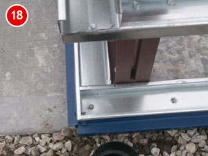 Close-up of a steel building frame corner on a concrete foundation, with a partially visible number 18 in a red circle. The frame is secured with screws, illustrating precision in construction. Pebbles are scattered on the ground in the lower part of the image.