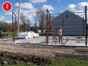 A construction site with a partially built steel structure on a concrete foundation. A person is working near a ladder, with building materials and a tractor nearby. A house and trees are visible in the background under a blue sky, showcasing the art of steel building construction.