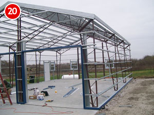 A partially constructed steel frame of a building stands on a concrete base. Construction tools and materials are scattered around the site. The number 20 is displayed in a red circle in the top left corner, highlighting this stage of construction.
