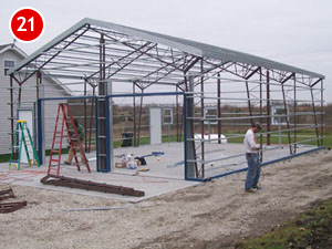 A partially constructed steel building framework with workers and ladders surrounds it. The structure stands on a concrete base in an open area beside a house, typical of modern steel building construction. Various construction materials are scattered on the ground.