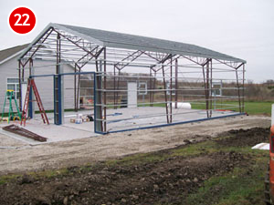 A partially constructed steel building with a roof appears amidst scattered tools and a lone ladder, resting on the bare ground. In the background, a completed structure stands proudly. A red circular number "22" marks the top left corner.