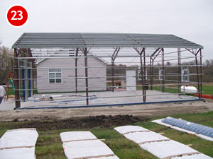 A partially constructed steel-framed building stands on a concrete foundation. A house is visible in the background. Construction materials are laid on the grass in the foreground, depicting active steel building construction. A red circle with the number 23 is in the top left corner.