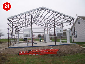 A steel building framework under construction stands on a concrete foundation, with workers visible in the background. A red ladder lies on the ground in the foreground, while a house is partially visible to the right.