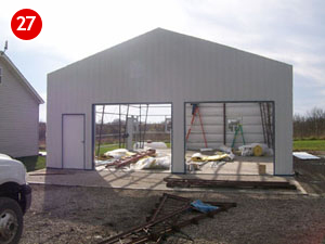 A partially constructed steel building garage with an open front is shown. Construction materials and ladders are inside. A white truck is partially visible on the left, and a small building is on the far left. A red number "27" is in the top left corner.