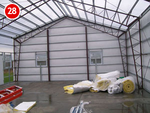 The interior view showcases a steel building construction with metal-framed structures and clear roofing. Two windows are visible, while construction materials, including insulation rolls and a ladder, are scattered on the floor. A red number "28" is prominently displayed in the top left corner.