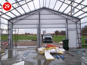The partially constructed steel building frame, with its clear roof, offers a view from inside. A truck is parked in the open area, surrounded by construction materials scattered on the concrete floor. In the background, a grassy field stretches beyond the scene of ongoing building efforts.