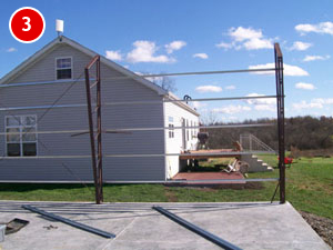 A partially constructed steel building frame stands on a concrete slab in front of a white house with a sloped roof, surrounded by grass. A wooden deck extends from the house, and the sky is partly cloudy.