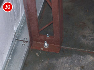 A steel frame is bolted to a concrete floor, indicative of building construction. The brown structure rests on a rectangular base plate with visible bolts. A red circular label with the number 30 adorns the top left corner, while the dark floor beneath appears slightly reflective.
