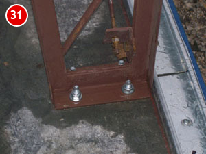 A metal bracket, essential for steel building construction, is secured by two bolts on a concrete surface, next to a metal edge. A red circle with the number 31 adorns the top left corner.