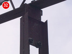 A close-up view of a rusty metal beam with two bolts, integral to steel building construction, positioned vertically under a horizontal beam against a cloudy sky. A red circle with the number 32 sits in the top left corner.