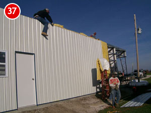 Two people are working on a steel building. One man is climbing down a tall ladder, while another stands near the base holding a tool. The sky is clear, and there is construction material on the rooftop, signaling ongoing construction efforts.