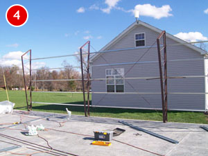 A gray house with a small window stands in the background. In the foreground, steel frames and construction materials are laid out on unfinished concrete. The clear sky, dotted with a few clouds, complements the setting. A red circle with the number 4 is in the top left corner.