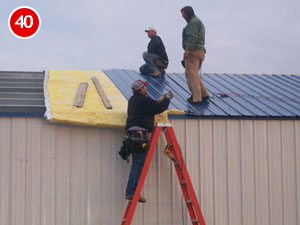 Three workers are engaged in steel building construction, installing a blue metal roof. One is on a red ladder holding a tool, while the others stand on the roof. Yellow insulation material peeks through. A red number 40 marks the top left corner.