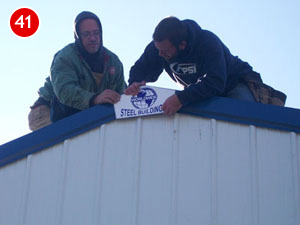 Two men in hoodies are busy with steel building construction, placing a "Steel Building" label atop the structure. The sky is clear and blue, with a bright red number "41" marking the upper left corner of the image.