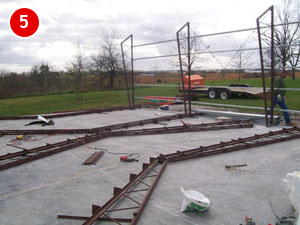 A construction site with a partially built steel framework on a concrete foundation. Metal beams and tools are scattered around. In the background, there is a grassy field and a trailer. The sky is cloudy. A red number "5" is in the top left corner, marking the steel building construction progress.