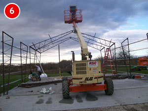 A construction site showcases a partly constructed steel building frame. A yellow lift with a worker in a basket rises towards the top of the structure. Under the cloudy sky, tools and materials are scattered around, embodying the progress of modern steel construction.