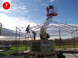Two workers on a construction site are using a lift to assemble a metal framework for a steel building. One worker is on the lift's platform, while the other climbs the structure. The sky is partly cloudy, and a vehicle is parked nearby.