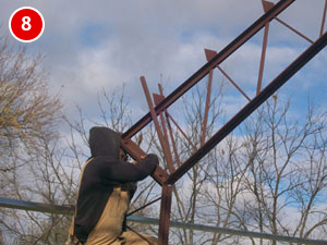 A person in a hooded jacket and overalls is climbing a metal ladder on a steel building construction site. Leafless trees and a partly cloudy sky serve as the backdrop. A red circle with the number 8 is in the upper left corner.