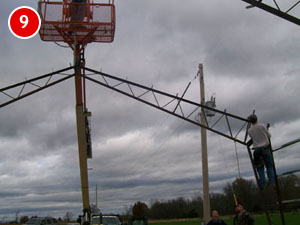 Two workers are engaged in steel building construction, installing a structure with a crane lift on a cloudy day. One stands on a boom lift basket, while the other remains grounded in safety gear. The structure seems to be a metal truss or framework.
