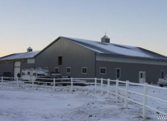 A large, gray barn with a snow-covered roof sits behind a white fence. Nearby, several vehicles and equipment rest under clear skies suggesting a cold winter day. Adjacent to the barn, metal garage kits offer seamless storage for an array of tools and machinery.
