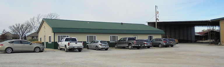 A one-story building with a green roof and beige siding sits amidst parked cars on a gravel lot. An open garage or warehouse space, possibly from Worldwide Steel, is attached to the right side. The overcast sky looms in the background, highlighting its robust construction by a renowned manufacturer.