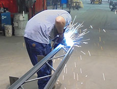 In a spacious industrial workshop, a person is welding a metal framework, with sparks flying as they skillfully join sections for Worldwide Steel. Dressed in protective gear, including a welding mask and gloves, they work amidst various tools and equipment essential for crafting durable steel buildings.