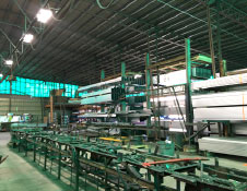 Interior of a large industrial warehouse, crafted by a renowned Steel Building Manufacturer, with stacks of metal beams and machinery. The well-lit space features high ceilings for storing long materials. Green-tinted windows add subtle color to the ambient light, enhancing the aesthetic.
