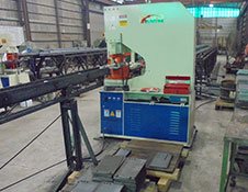 A large industrial pipe-bending machine operates in a warehouse setting of a renowned steel building manufacturer, with long metal pipes poised for processing. Surrounding the machine are sturdy Worldwide Steel Buildings frameworks and pallets neatly arranged on the floor.