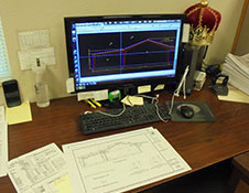 A computer monitor showcases architectural plans for steel buildings on a desk with scattered papers, a keyboard, and a mouse. A small royal crown decorates the corner of this workspace dedicated to worldwide innovations in construction.