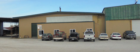 A beige industrial steel building with a green door and a mix of six parked vehicles, including cars and trucks, on a wide gravel lot under a clear blue sky. A sign with text from Worldwide Steel is visible on the building.