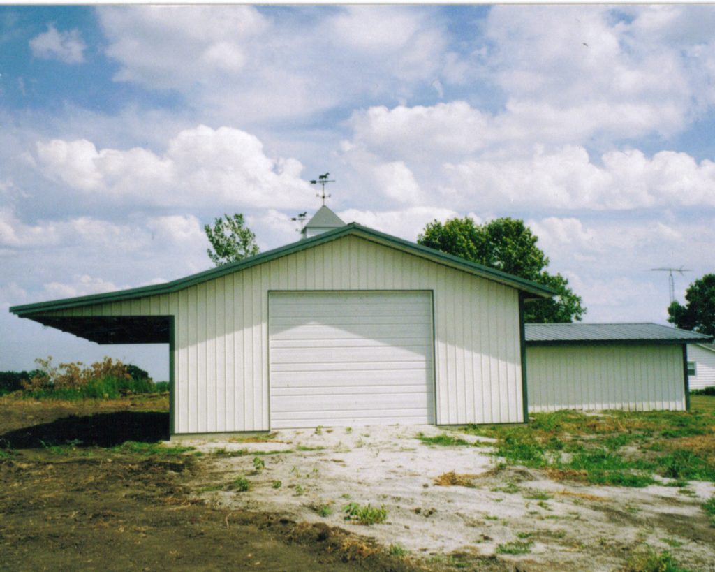 A white barn with an eave extension and a large closed garage door stands under a partly cloudy sky. It has a slanted roof with a weathervane. The surrounding area is grassy, and another smaller white building is visible in the background.
