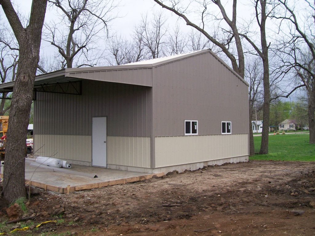A large, partially constructed metal building stands in a clearing surrounded by leafless trees. The structure features a slanted roof with eave extensions, a closed door, and windows. The ground is cleared, with dirt and a concrete slab visible.