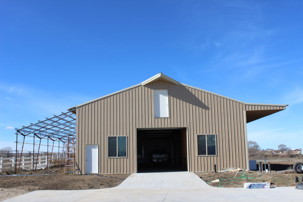 A large, tan metal barn under a clear blue sky features an open sliding door, revealing a car inside. An eave extension from one side adds architectural flair. The ground is bare with some construction materials nearby.