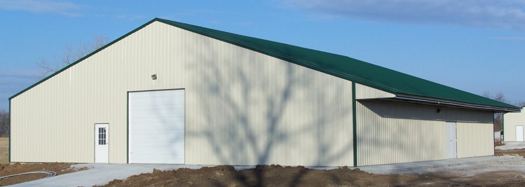 A large beige metal building with a green roof and a prominent eave extension features a small door and a large garage door. It stands proudly against a clear blue sky, with the shadow of a tree cast gracefully on its side.