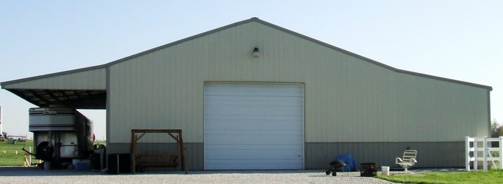 A large metal barn with a closed white garage door sits under an eave extension, perfectly shading the gravel driveway. To the left, there's an open section with a horse trailer and a wooden bench, while on the right, some outdoor furniture and equipment are visible.