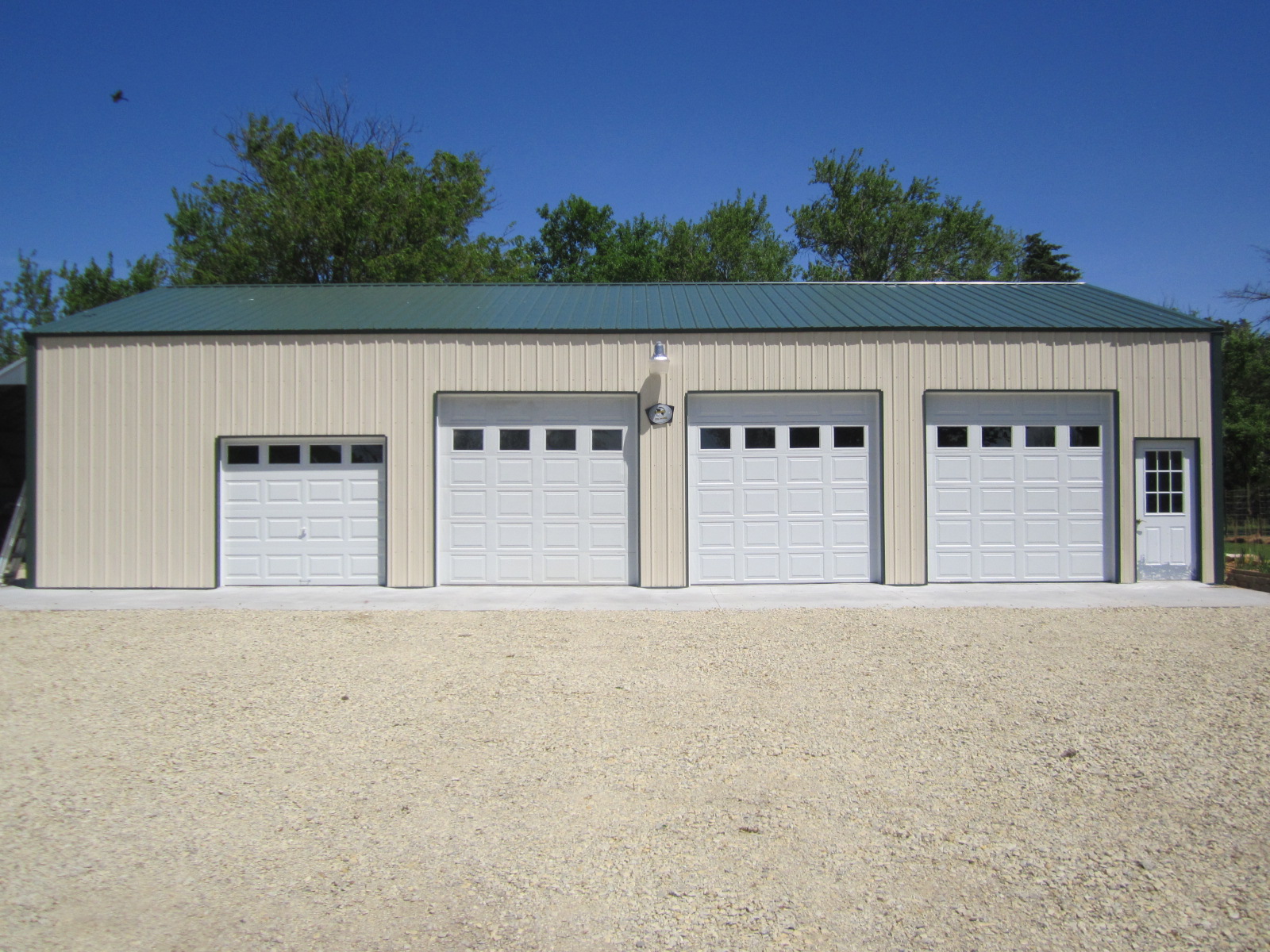 The exterior showcases a beige metal garage with a green roof and four white roll-up doors, exemplifying a seamless steel building process. Nestled on a gravel lot with trees and a clear blue sky, it promises an exceptional client experience from start to finish.