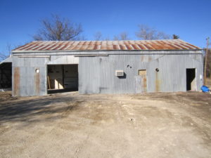 A weathered metal shed with a rusty roof stands on a dirt lot, illustrating the real-life experience of enduring elements. The front features two large garage doors, and sparse trees can be seen in the background under a clear blue sky.