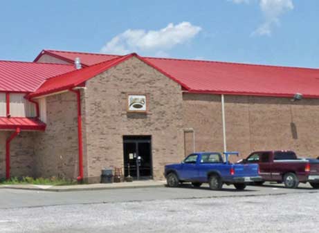 A large brick building with a red metal roof stands under a blue sky. Two pickup trucks, one blue and one maroon, are parked in front of the entrance, displaying its affordable style. This inviting structure hints at its versatile purpose, possibly housing steel garages within.