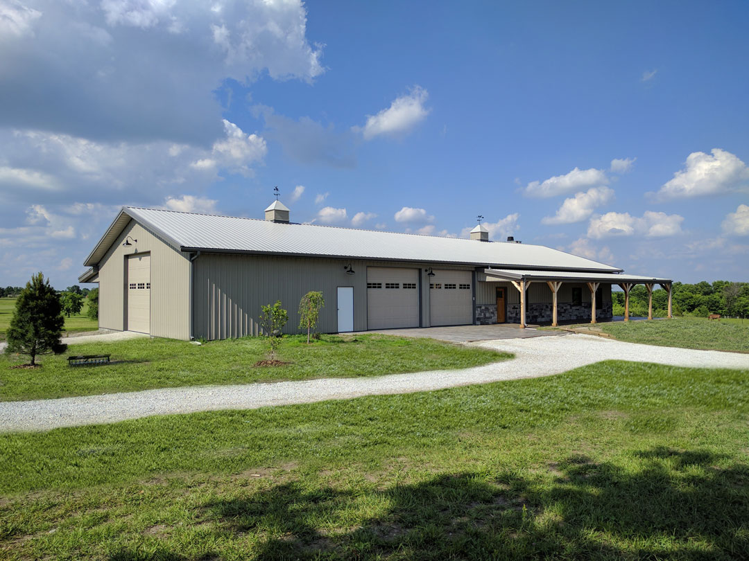 A large beige metal barn with a peaked roof, perfect for an AWS Certified Welding Fabricator, stands under a partly cloudy sky. It features multiple large garage doors and a covered porch area. A gravel path leads to the structure, surrounded by green grass and a couple of small trees.
