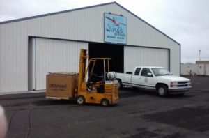 A forklift carrying a large box marked "Dynasol" moves in front of a white industrial building with a sign that reads "Wings of Faith Ministries.” Nearby, a white pickup truck is parked, underscoring the seamless operation crucial for customer success.