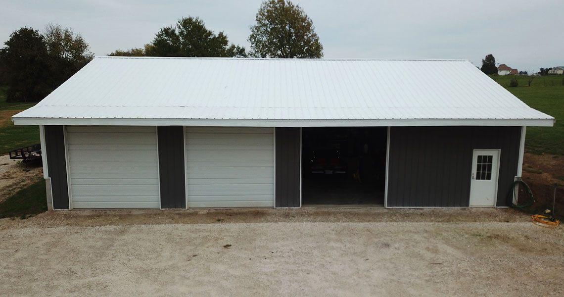 A large, white-roofed metal building with three garage doors and a small entry door on the right. It is situated on a grassy landscape with a few trees in the background. The ground in front of the building is gravel.